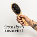 Hand håller en Green Heads hårborste mot vit bakgrund. Texten Green Heads borstmetod syns i bild.
